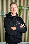Lächelnder Mann im schwarzen Hoodie mit verschränkten Armen vor einer gestreiften Wand.