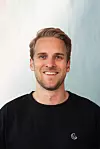 Lächelnder Mann im schwarzen T-Shirt vor hellem, neutralem Hintergrund.