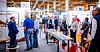 Menschen stehen auf einer Fachmesse an einem Stand mit technischen Exponaten und Prospekten.